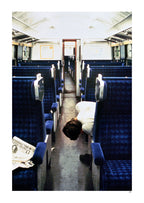 Person lying on a train seat with blue seats and a newspaper on the floor.